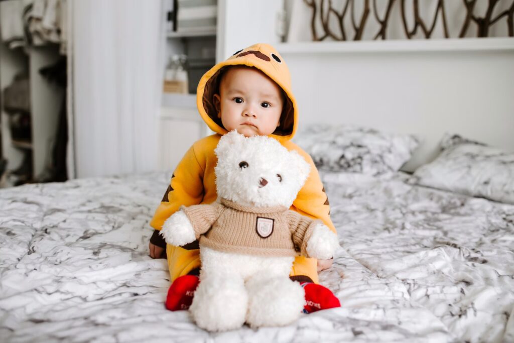 foto eines babies mit gelbem hoodie und stoffbären auf einem bett in die kamera schauend