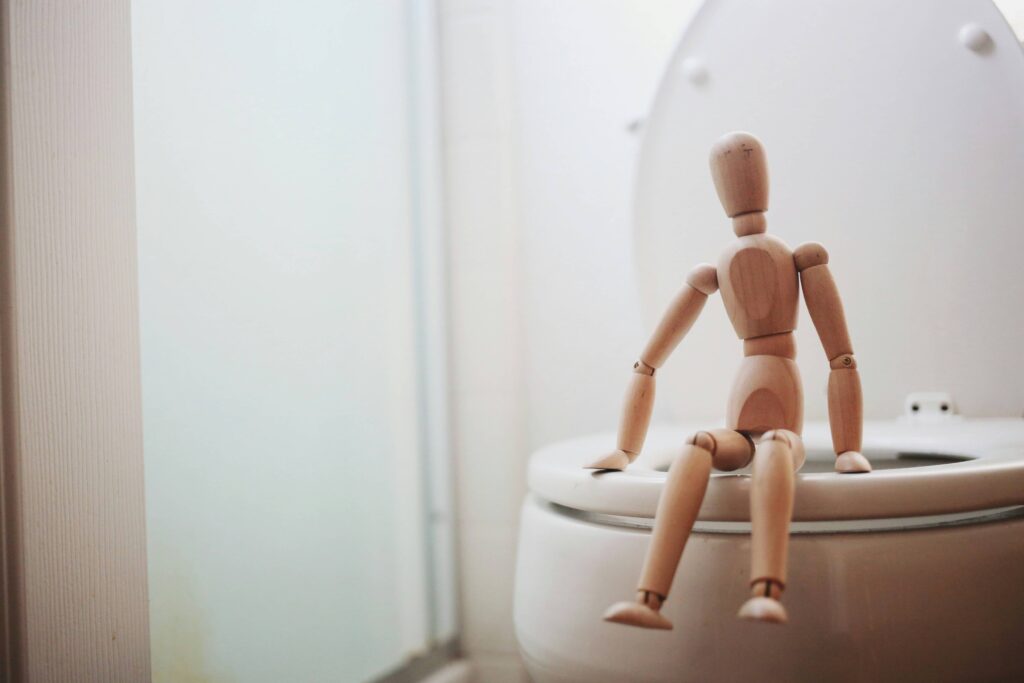 Eine hölzerne Gliederpuppe sitzt auf einer Toilette, symbolisch für das häufige Wasserlassen bei einem entgleisten Blutzuckerspiegel.