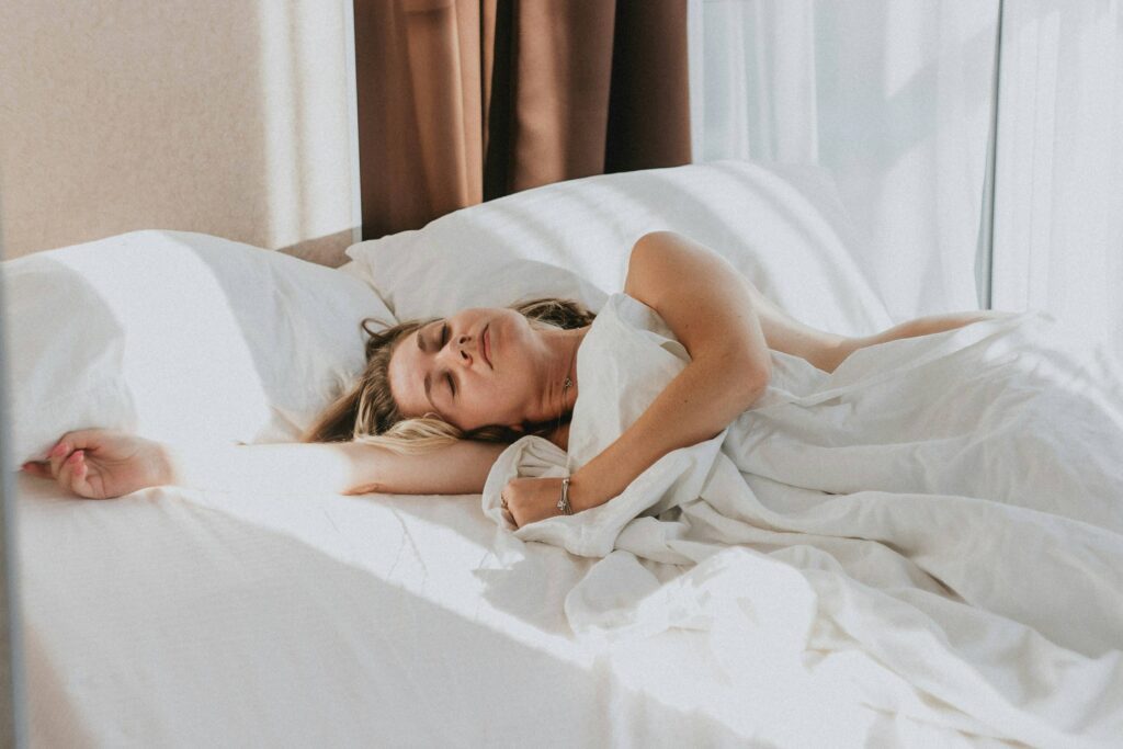Eine junge Frau schläft friedlich in einem sonnendurchfluteten, weißen Bett, was die wichtige Regeneration beim Abnehmen darstellt.