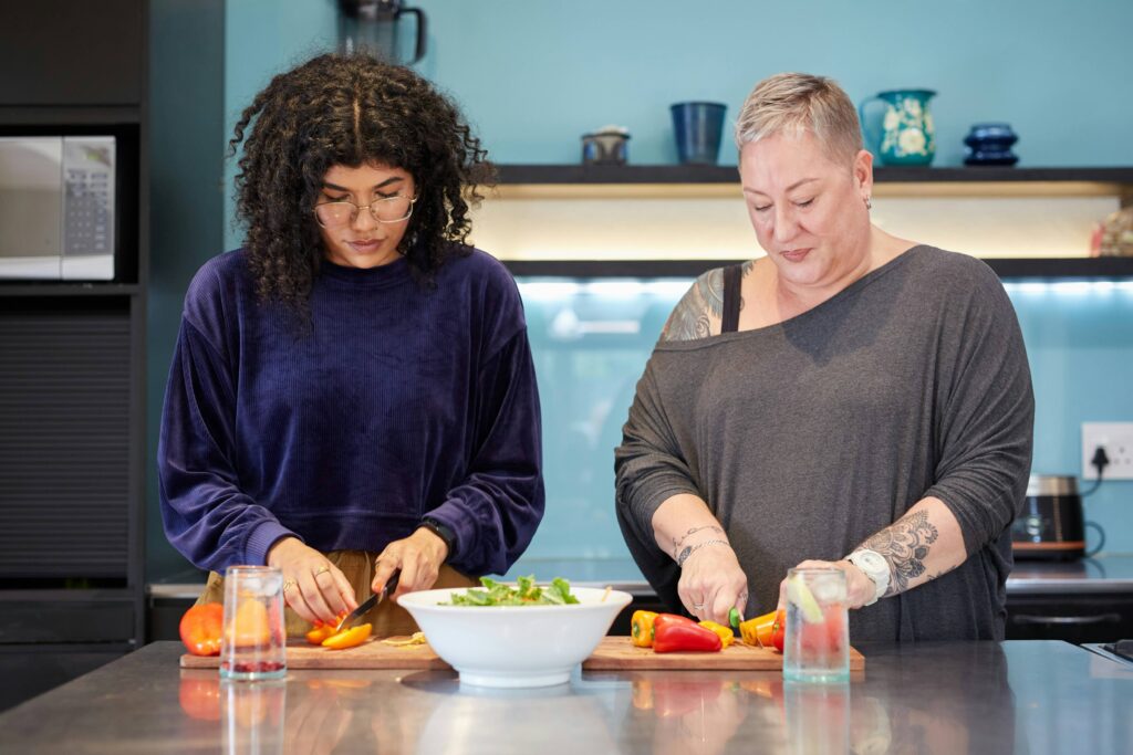 Zwei Frauen bereiten in einer Küche entspannt gemeinsam einen frischen Salat zu, was eine positive und alltagstaugliche Einstellung zur Ernährung symbolisiert.
