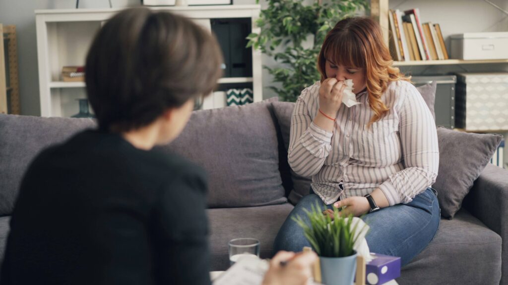 Eine weinende oder kranke Frau sitzt mit einem Taschentuch auf dem Sofa. Eine erhöhte Infektanfälligkeit zählt zu den typischen Frühzeichen